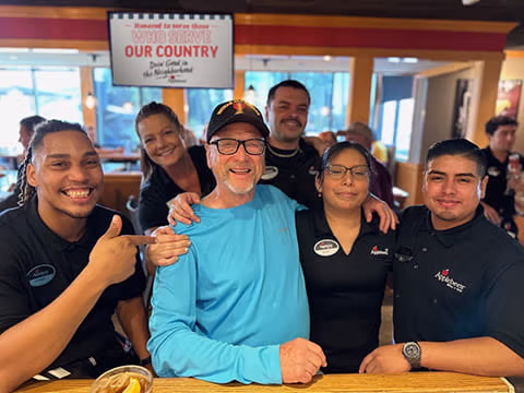 Applebee’s staff pose with a veteran in a blue shirt and cap seated at a table inside the restaurant